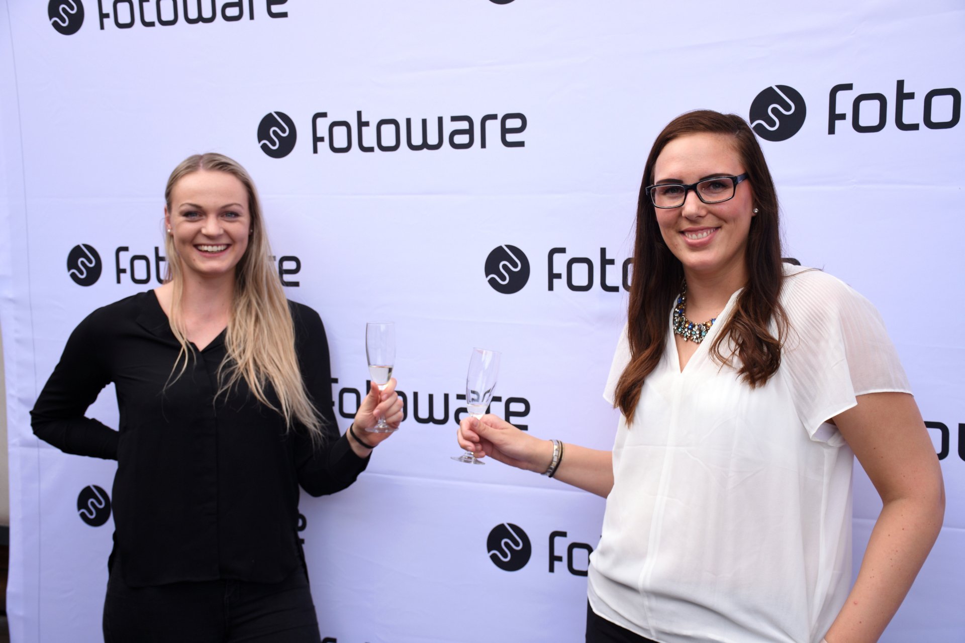 Two women smiling and holding glasses in front of a FotoWare logo wall